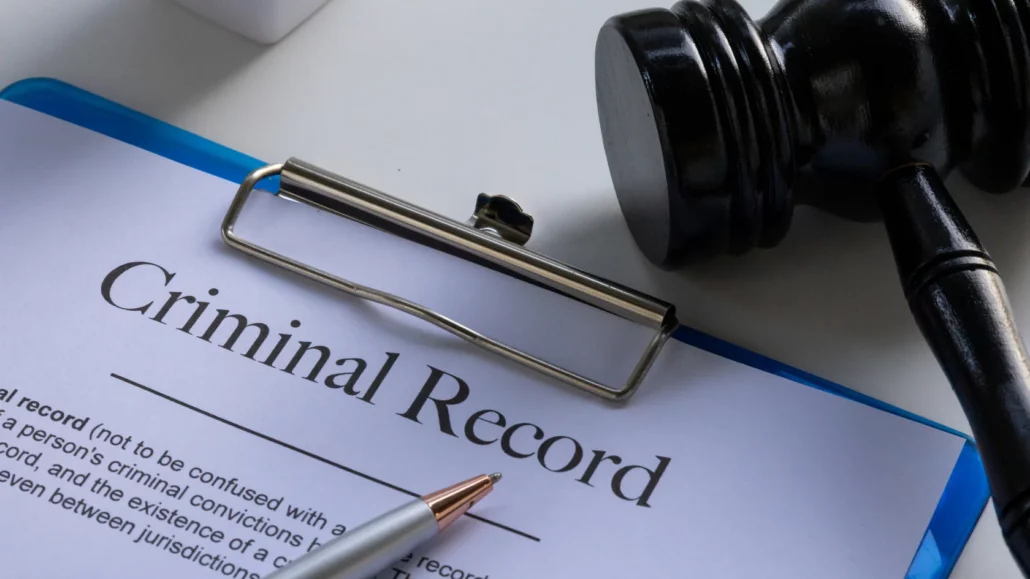 A close-up of a clipboard with a document titled Criminal Record, a silver pen, and a judge’s gavel on a white surface.