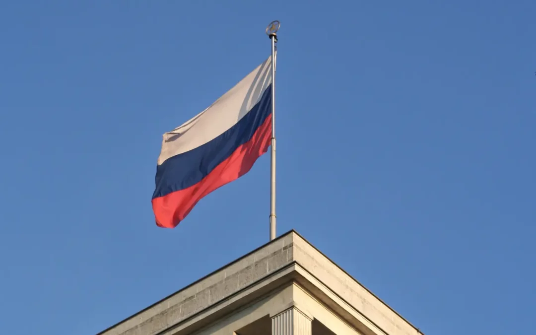A Russian flag with white, blue, and red horizontal stripes waves atop a building against a clear blue sky.