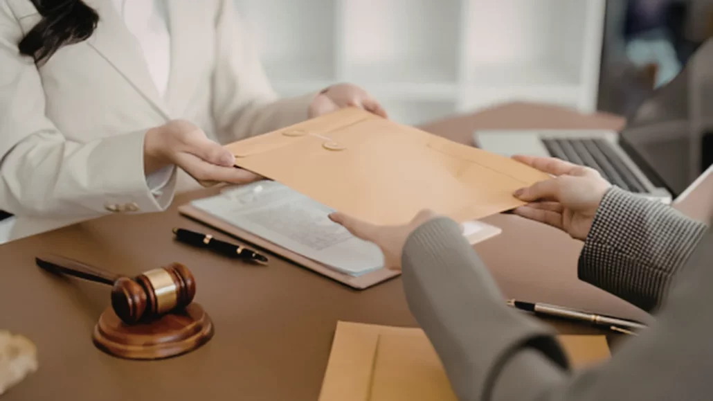 Two people exchange a large brown envelope across a desk with legal documents, a gavel, pens, and a laptop. Only their hands and part of their torsos are visible.