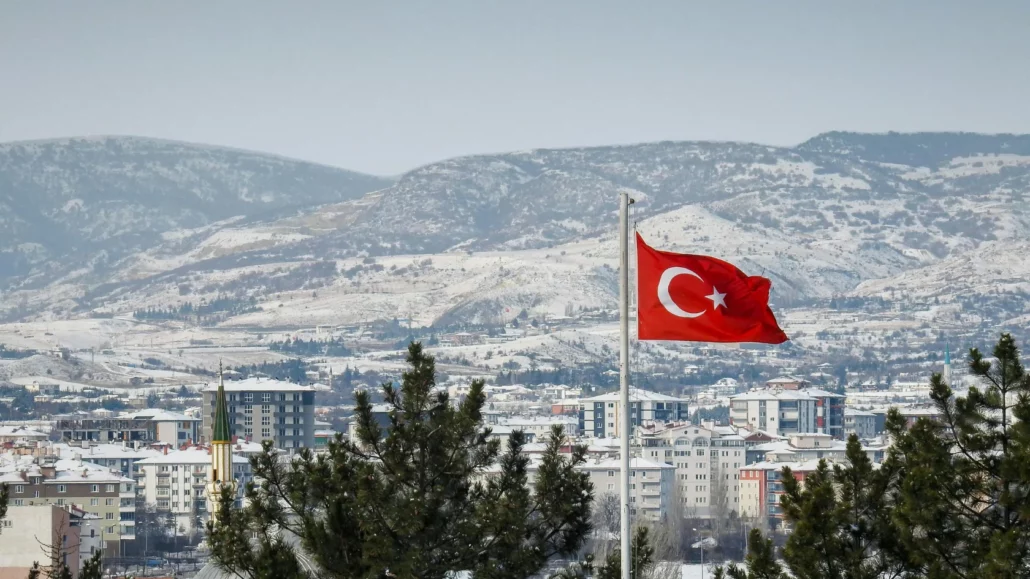 A Turkish flag waves on a flagpole in the foreground with snow-covered buildings and mountains in the background, framed by pine trees.