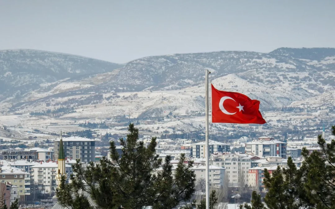 A Turkish flag waves on a flagpole in the foreground with snow-covered buildings and mountains in the background, framed by pine trees.