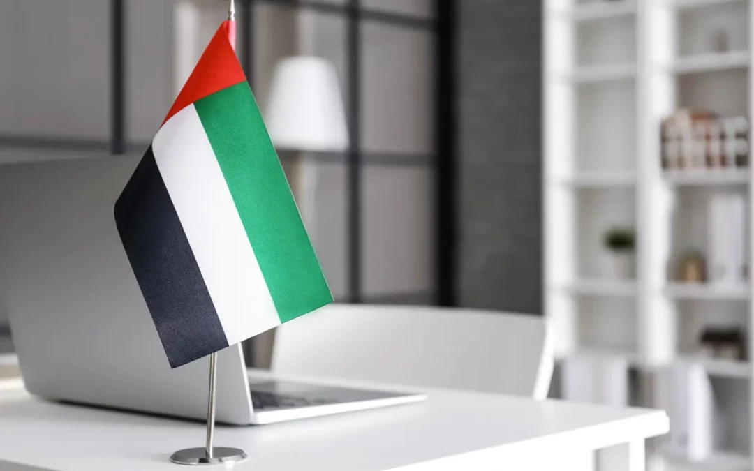A small United Arab Emirates flag stands on a white desk next to a closed laptop in a modern, bright office with shelves and a lamp in the background.