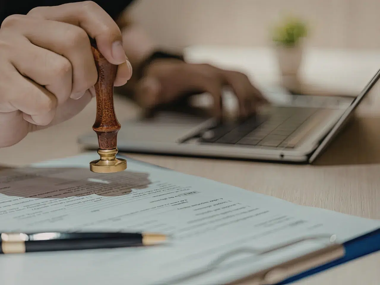 A person stamps a document on a desk, with a pen and clipboard nearby. In the background, another hand types on a laptop keyboard.