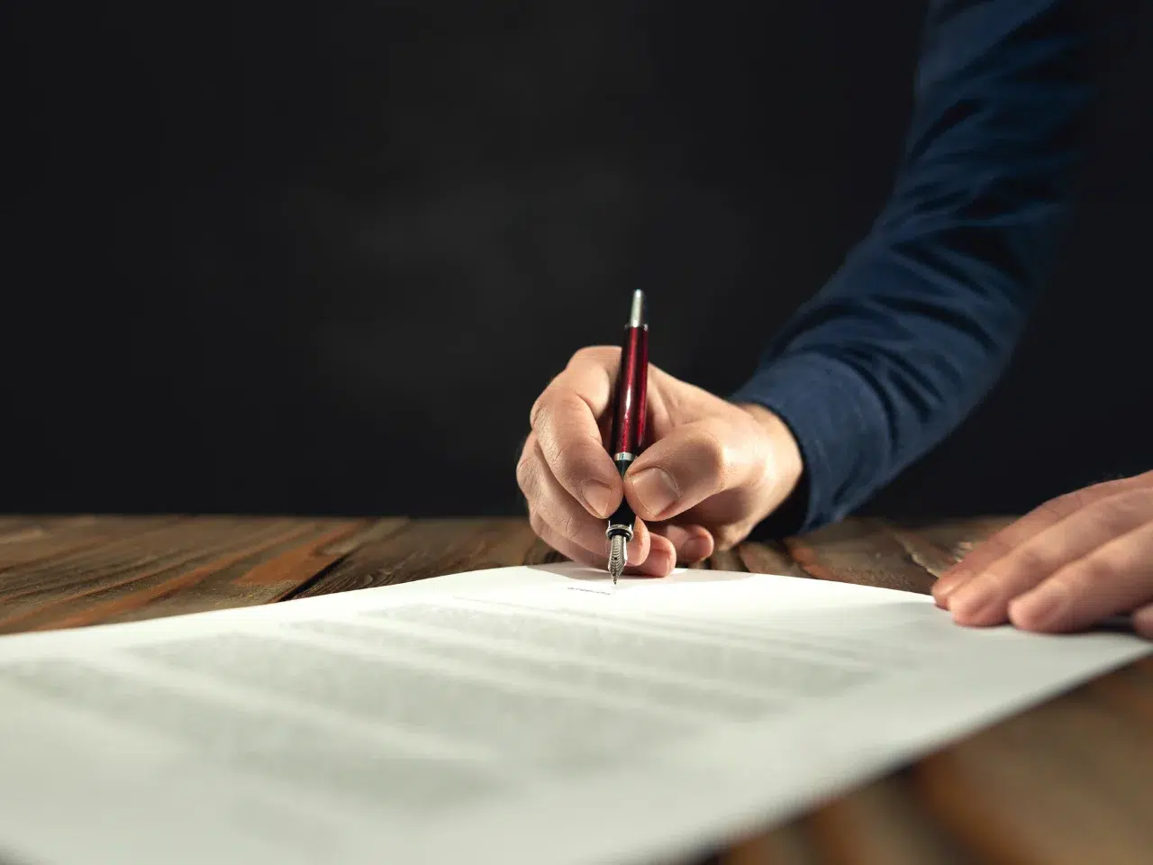 A person in a blue sleeve signs a document on a wooden table with a silver and red pen, with the background blurred and dark.