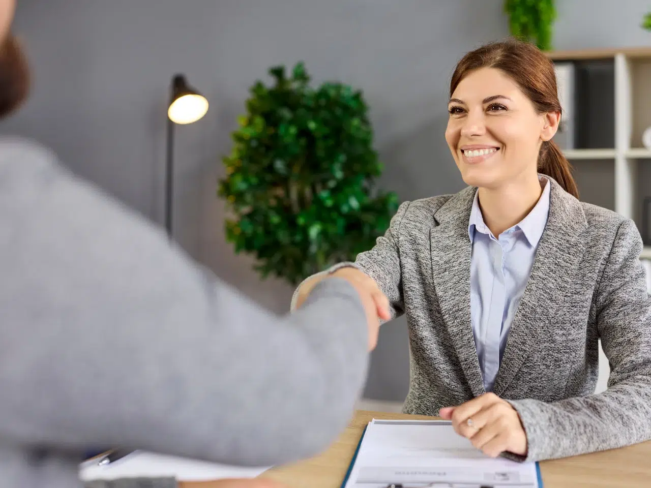 A woman in a blazer smiles and shakes hands with another person across a desk, suggesting a job interview or business meeting. A clipboard and pen are on the desk, and there is a plant in the background.