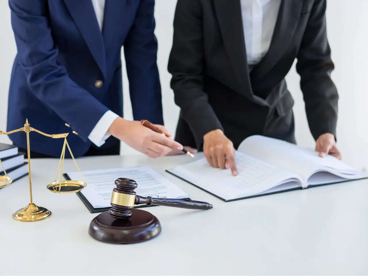 Two people in business suits review documents on a desk with a gavel and brass scales of justice, suggesting a legal or courtroom setting.