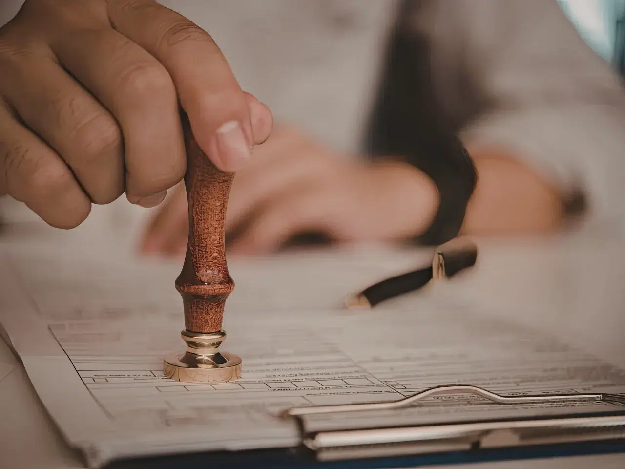 A person presses a wooden stamp onto a document on a clipboard, with a pen and blurred hand visible in the background, suggesting official approval or certification.