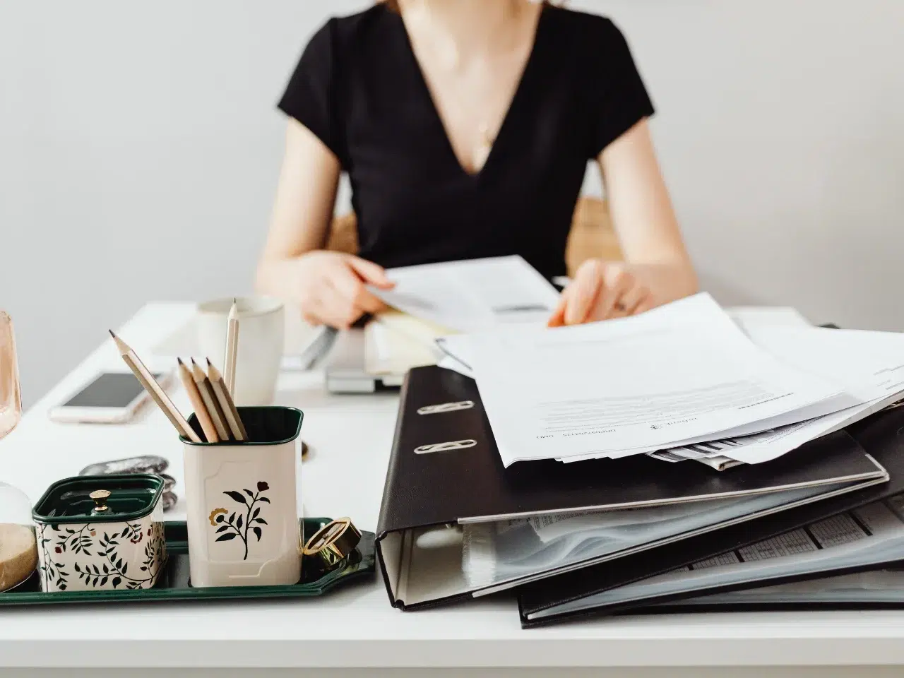 A woman in a black dress sits at a desk with folders, papers, a coffee cup, and a container of pens and pencils, appearing to review documents. Her face is not fully visible in the image.