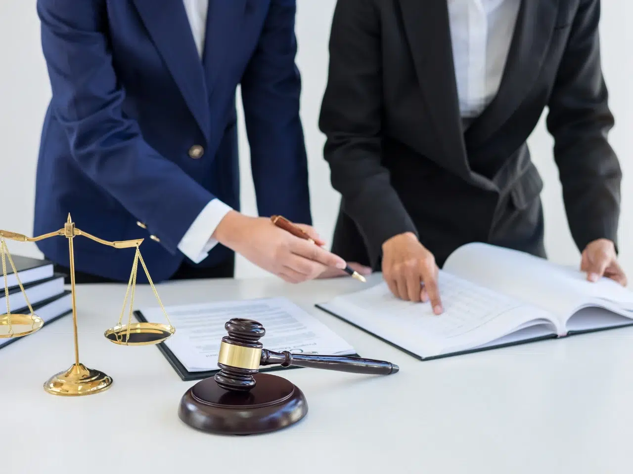 Two people in business suits stand at a desk reviewing documents. A gavel and scales of justice sit on the table, suggesting a legal setting or discussion. Neither person’s face is visible.