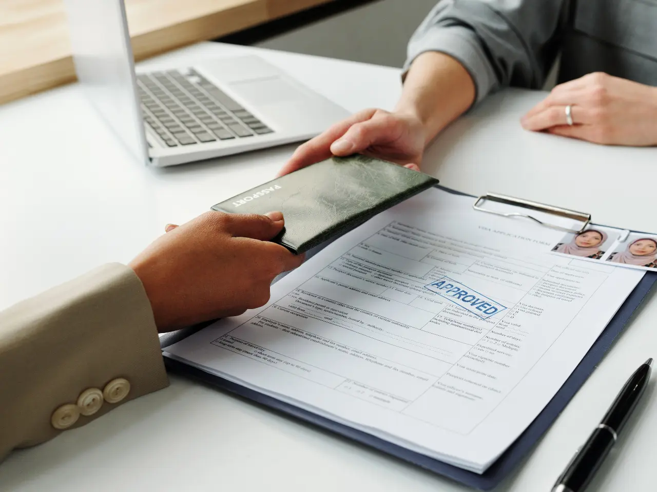 Two people sit at a desk with a laptop. One person hands a passport to another over a clipboard holding an approved visa application with passport photos attached.