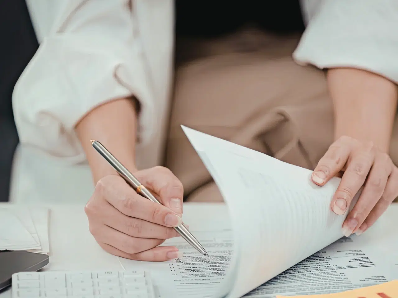 A person in a light-colored jacket holds a stack of papers with one hand and writes with a pen on a document with the other hand, sitting at a desk with documents and a calculator.