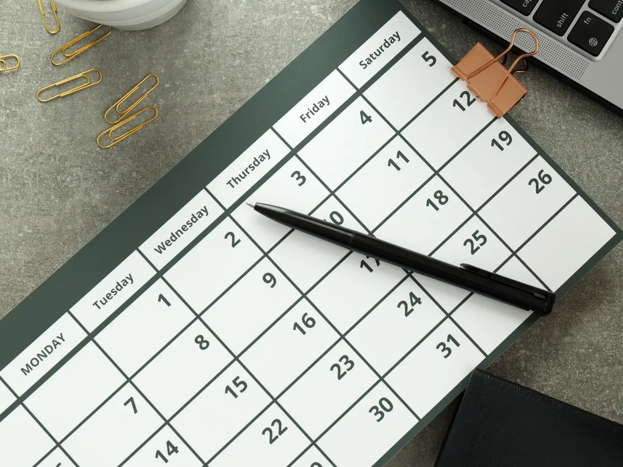 A black pen rests diagonally on a paper calendar displaying dates in a month. Paper clips, a binder clip, and part of a laptop are visible around the calendar on a gray desk.