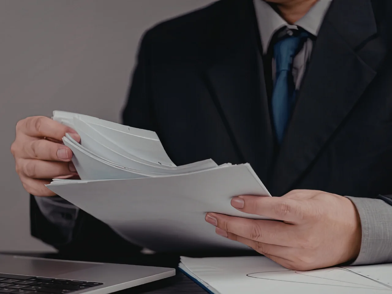 A person in a dark suit and blue tie is sitting at a desk, holding and reviewing a stack of documents beside an open laptop. The persons face is not visible.