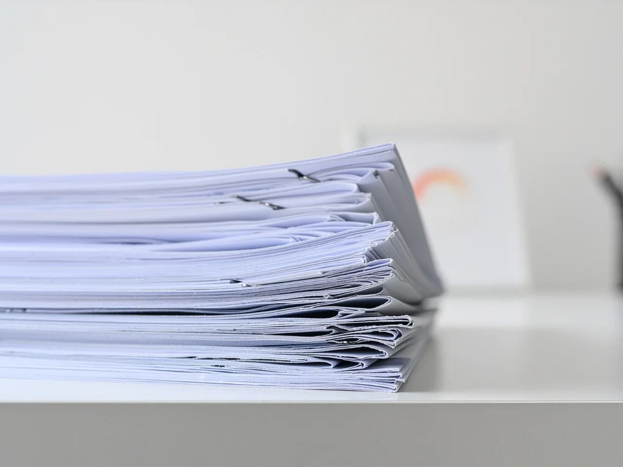 A close-up side view of two neat stacks of papers placed on a white desk, with a blurred background featuring a small object and a white item.