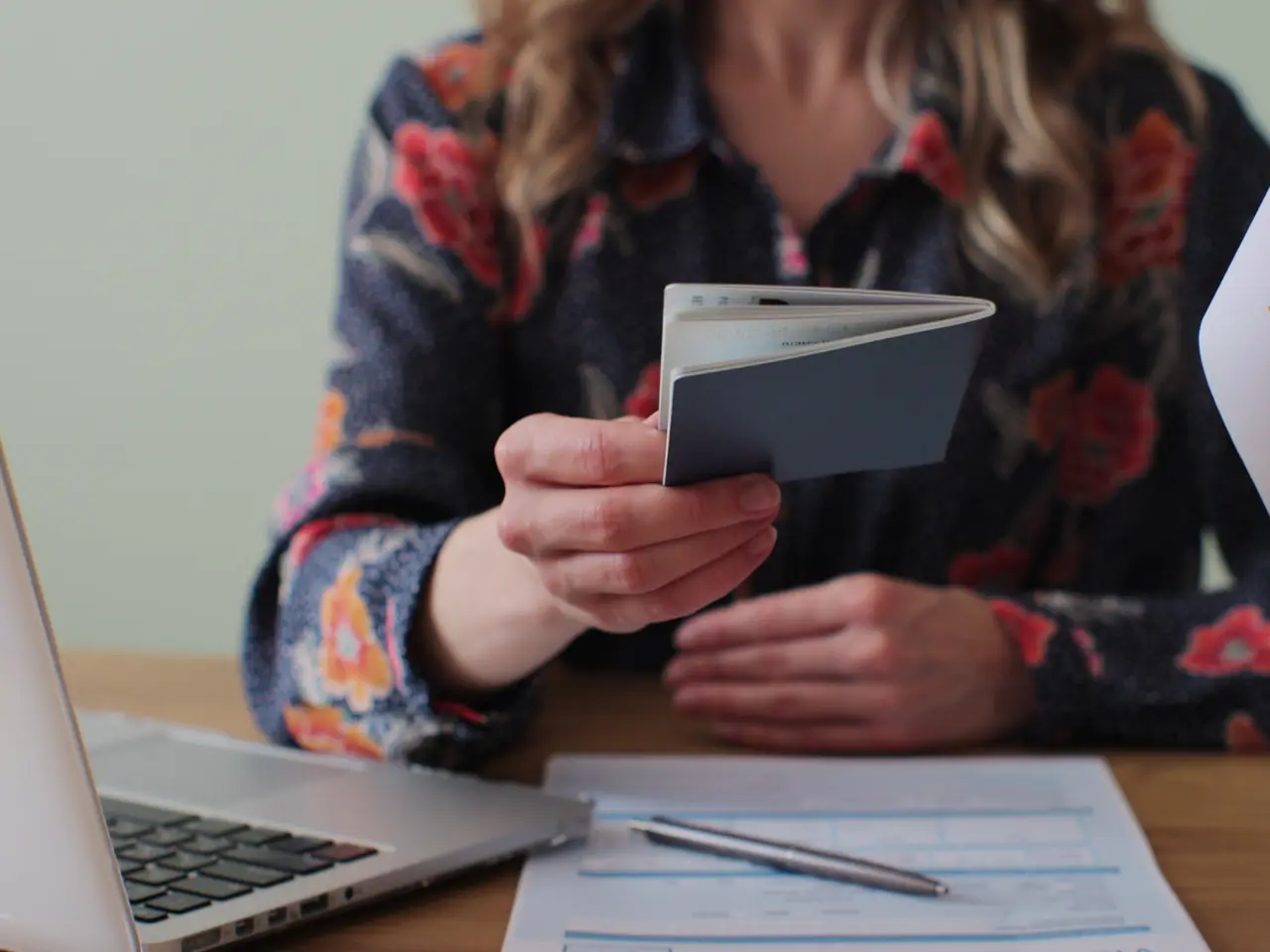 A person wearing a floral-patterned shirt sits at a desk, holding an open passport. On the desk are a laptop, a pen, and some documents. The persons face is not visible.