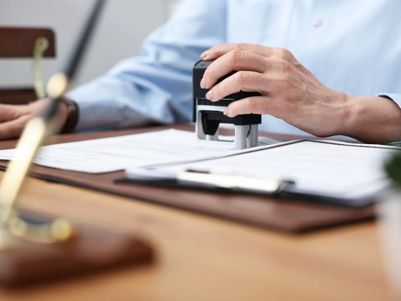 A person presses a stamp onto a document on a desk, with papers, a pen, and a decorative object nearby.