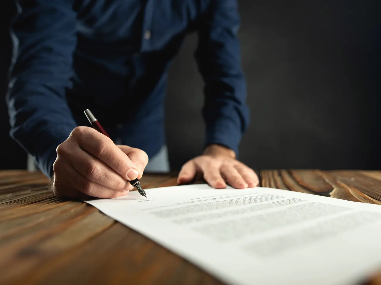 A person in a dark blue shirt is leaning over a wooden table, signing a document with a pen. The focus is on the hand holding the pen and the paper, while the background is blurred.