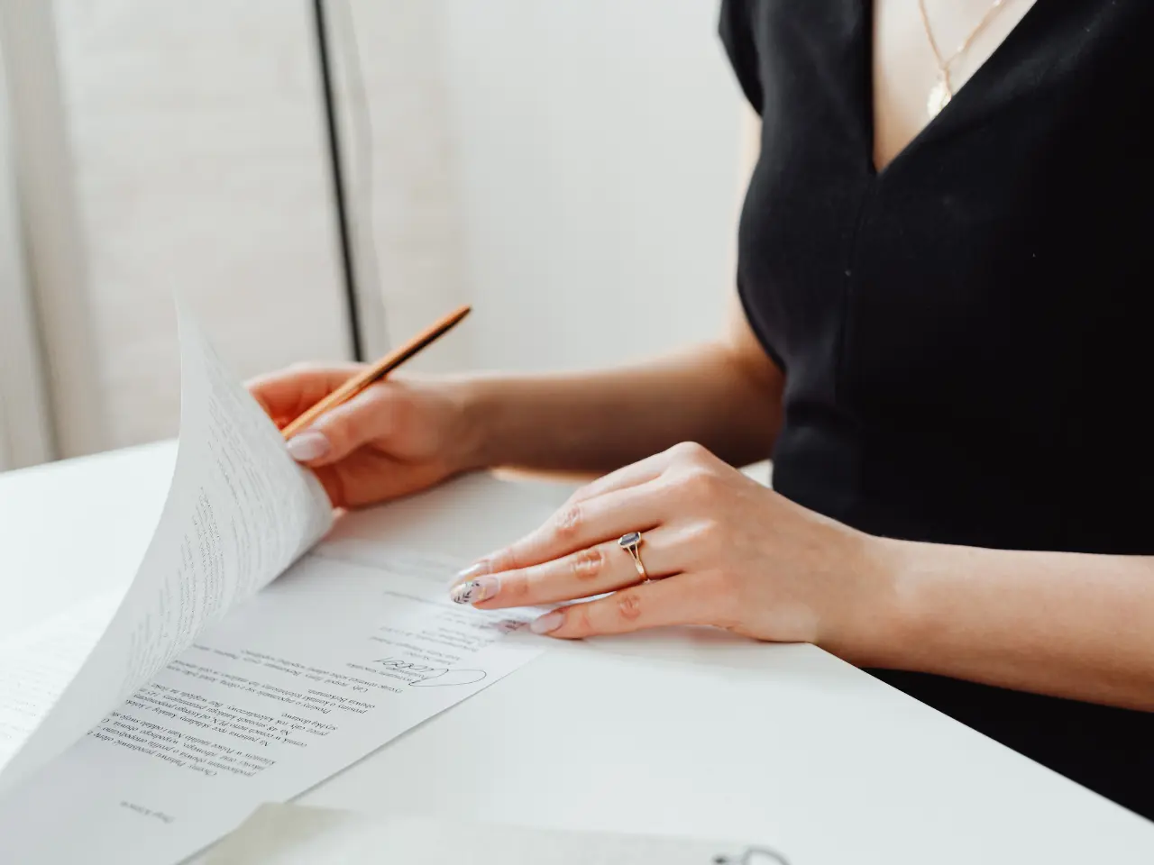 A woman in a black dress sits at a desk, holding a pencil and flipping through documents or papers, with only her torso and hands visible.