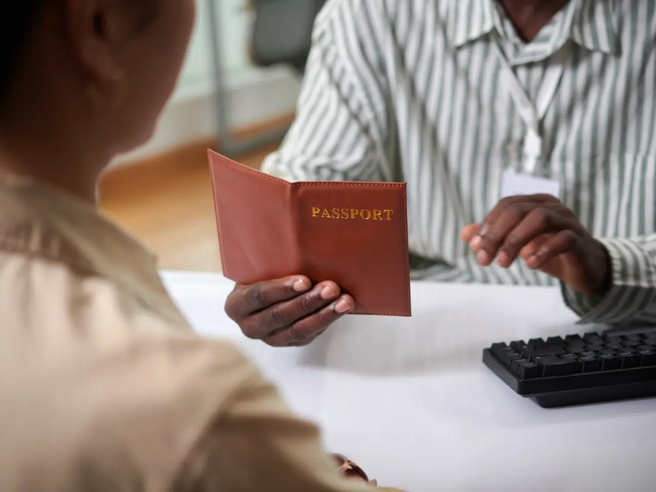 Two people sit across a desk; one holds an open brown passport while the other listens. A computer keyboard is on the desk, and both individuals faces are not fully visible.