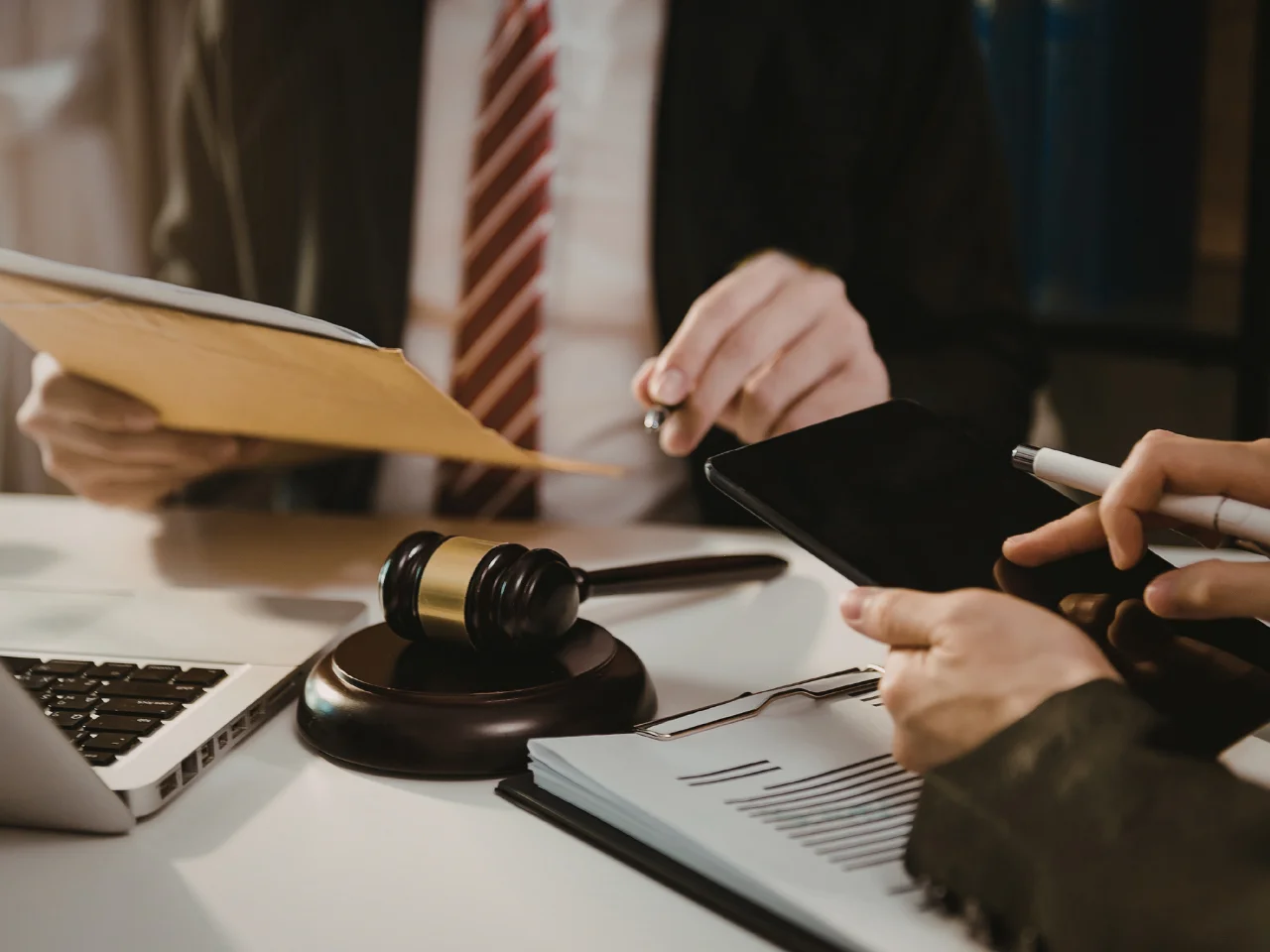 Two people in business attire sit at a desk with a gavel, tablet, documents, and a laptop, discussing legal paperwork in an office setting.