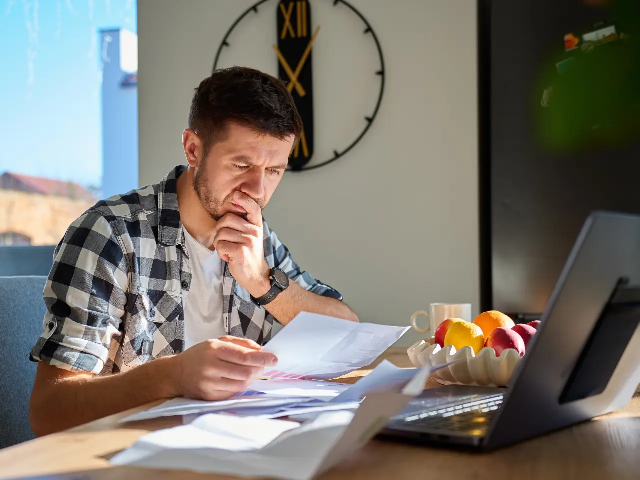 A man sits at a table covered with papers, looking thoughtfully at a document. A laptop, a bowl of fruit, and a coffee cup are on the table. A large wall clock is visible behind him. Sunlight shines through a window.