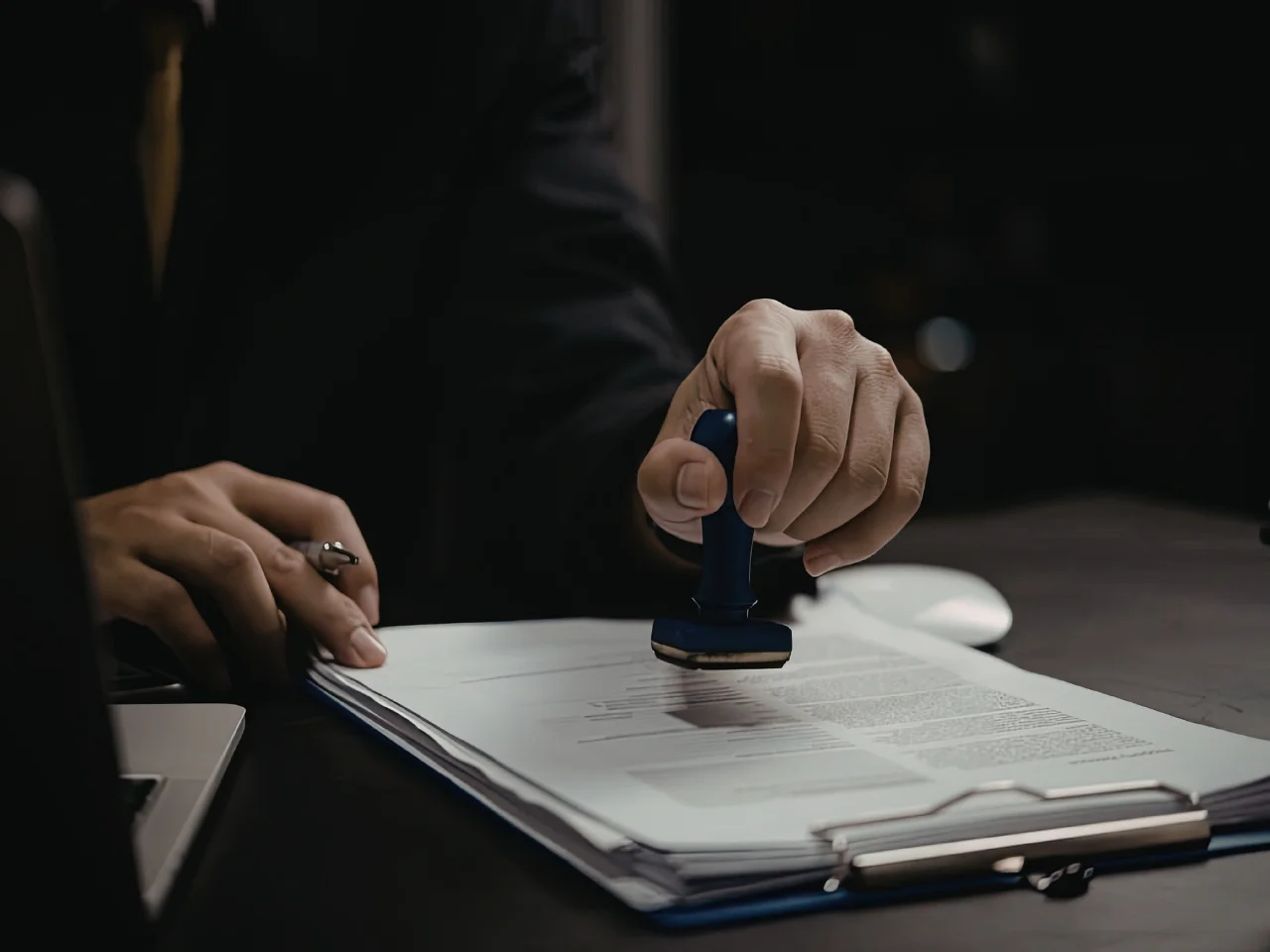 A person wearing a dark suit presses a blue stamp onto a stack of documents on a desk, with a pen and a laptop nearby.