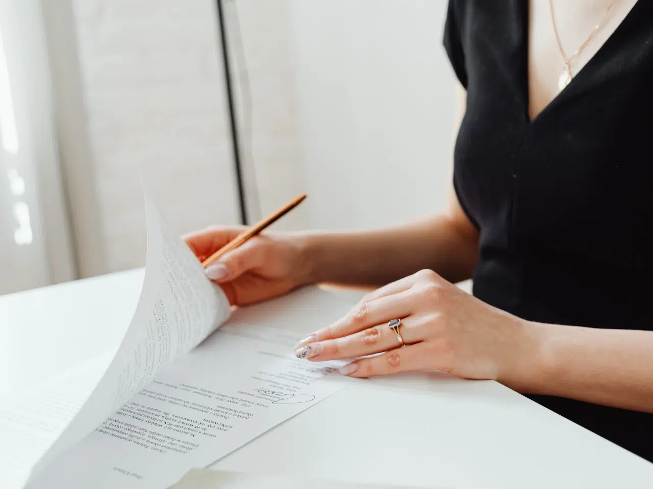 A person in a black shirt flips through documents on a desk while holding a pencil. Only their hands and part of their torso are visible. The scene is bright and minimalistic.