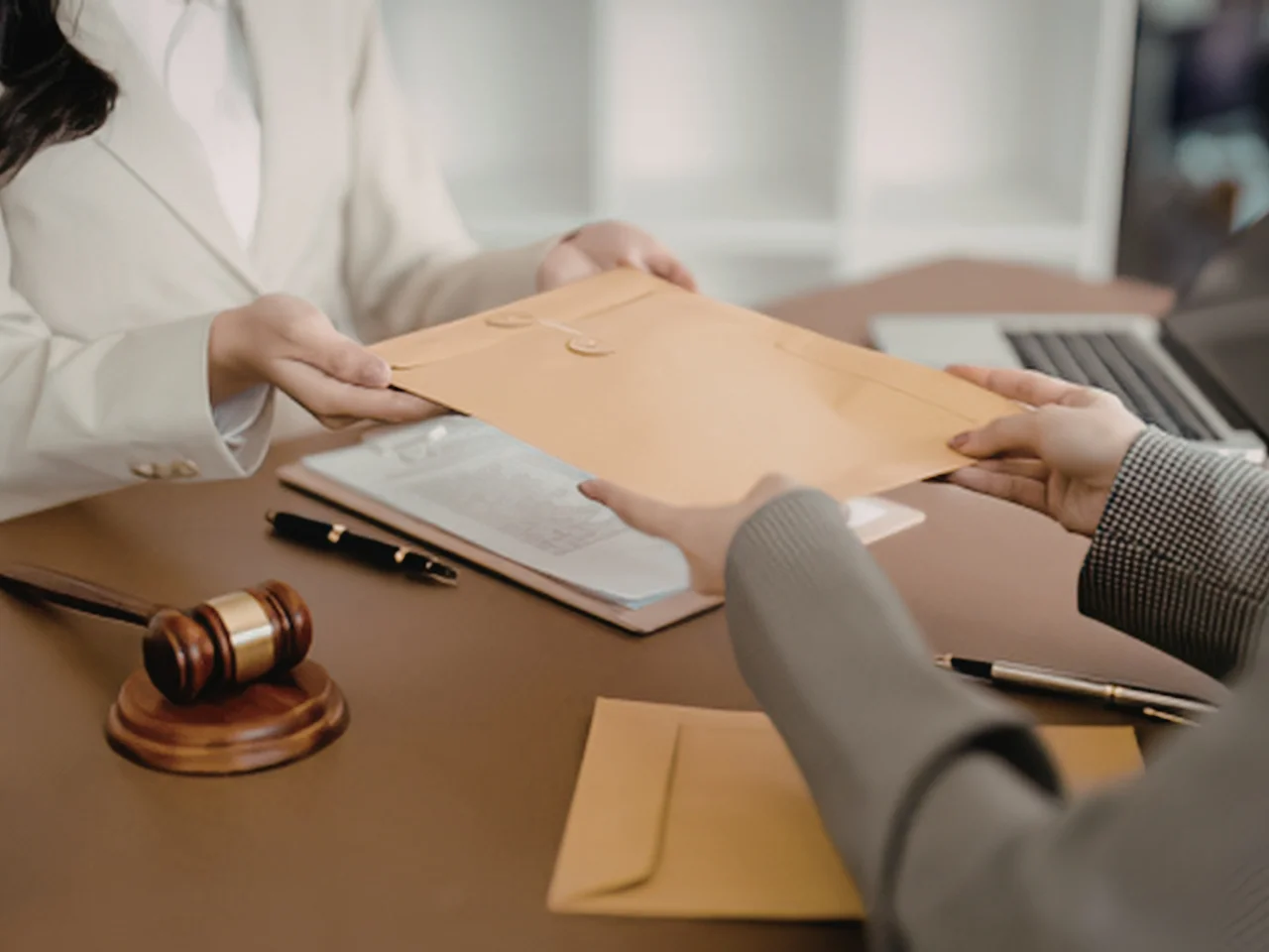 Two people in business attire exchange a large manila envelope across a desk with documents, a gavel, and a pen, suggesting a legal or official transaction.