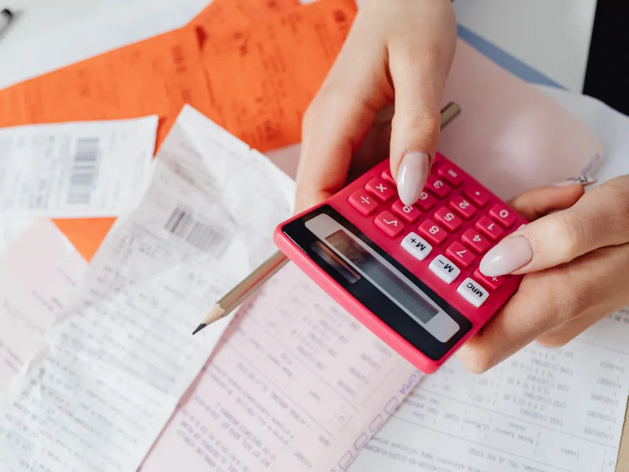 A person holding a pink calculator and a pencil, surrounded by receipts and orange bills on a desk, appearing to calculate expenses or manage finances.