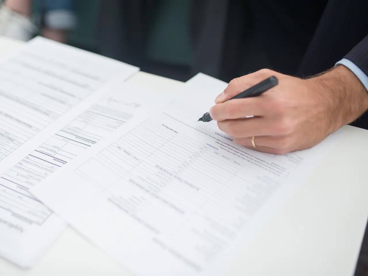 A person holding a pen fills out a form on a desk, with several other documents spread out nearby.