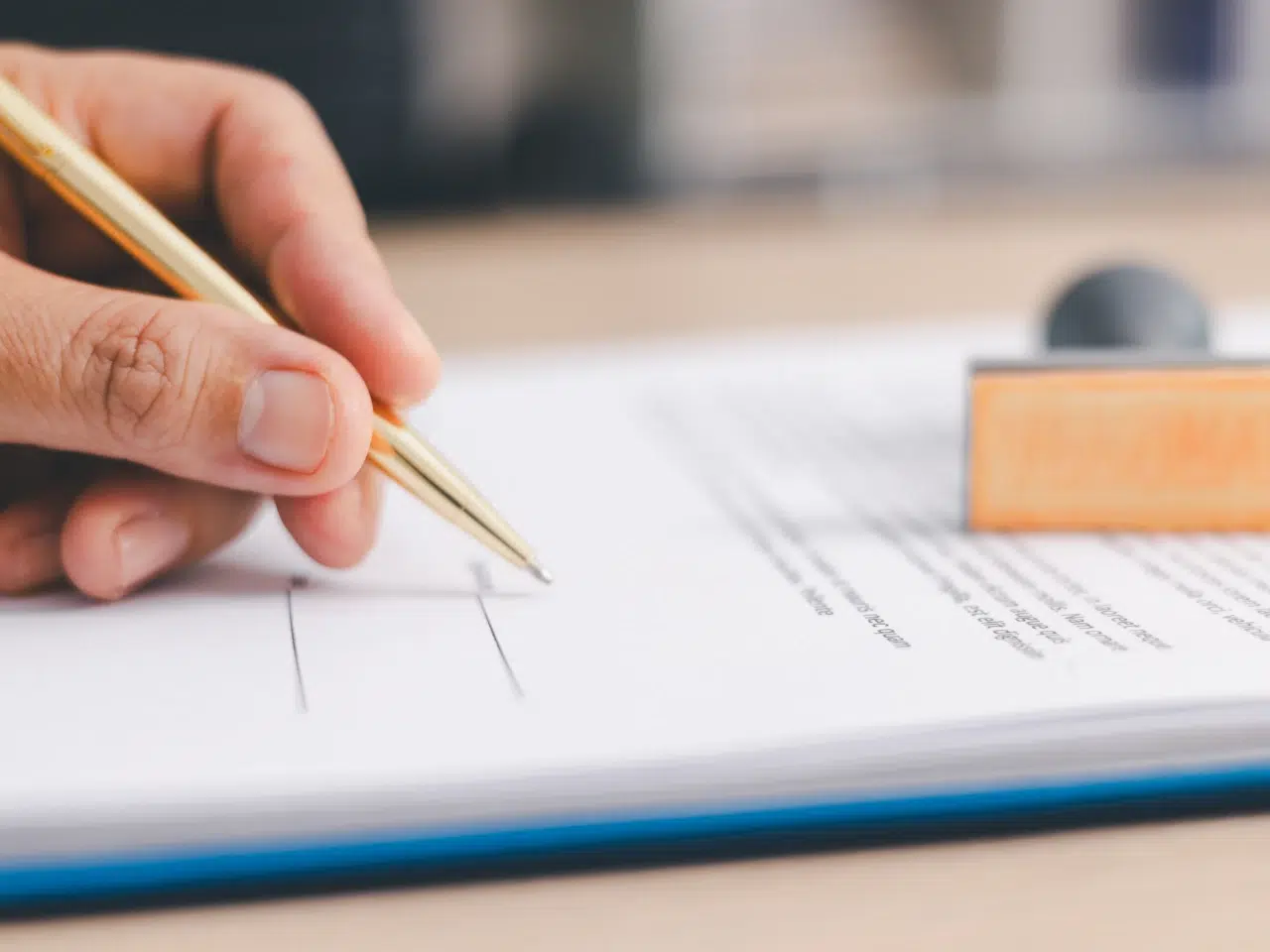 A close-up of a person’s hand holding a gold pen and about to sign a document on a desk, with an orange rectangular stamp visible next to the paper.