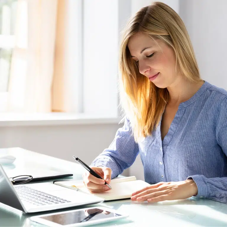 A woman with long blonde hair sits at a desk, writing in a notebook with a pen. An open laptop, tablet, and smartphone are on the desk beside her. She appears focused and is working in a bright, sunlit room.