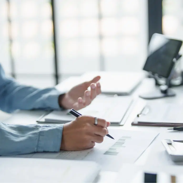 A person in a blue shirt holds a pen and gestures while reviewing printed charts at a desk with a tablet, papers, and documents in a bright office setting.