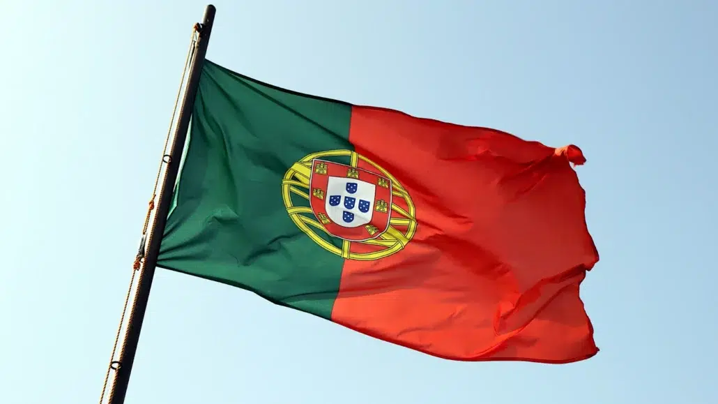 The flag of Portugal, with green and red vertical stripes and a coat of arms in the center, waves on a flagpole against a clear blue sky.