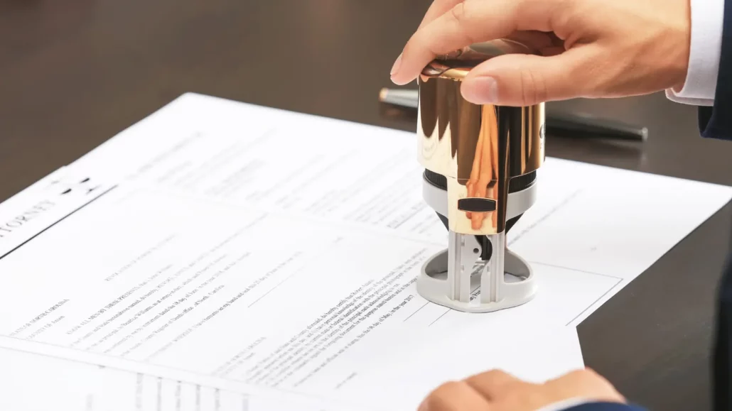 A person presses a gold-colored stamp onto a document, likely notarizing or certifying the paperwork on a dark desk. A pen lies nearby.