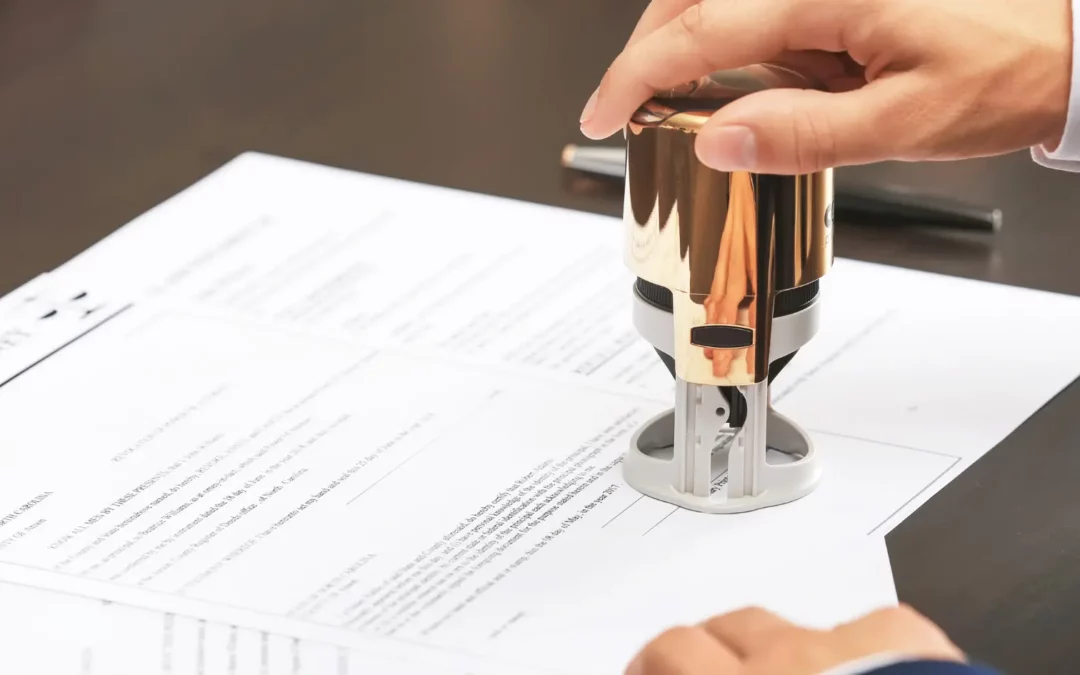 A person presses a gold-colored stamp onto a document, likely notarizing or certifying the paperwork on a dark desk. A pen lies nearby.
