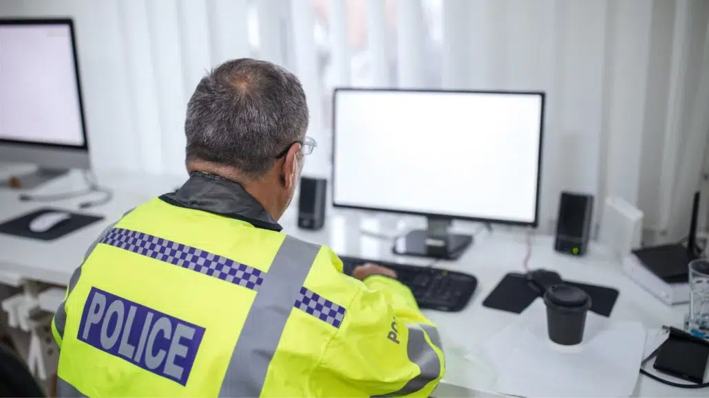 A police officer in a high-visibility jacket sits at a desk, working on a computer with a blank screen in a bright office setting.