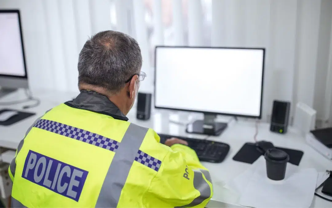 A police officer in a high-visibility jacket sits at a desk, working on a computer with a blank screen in a bright office setting.