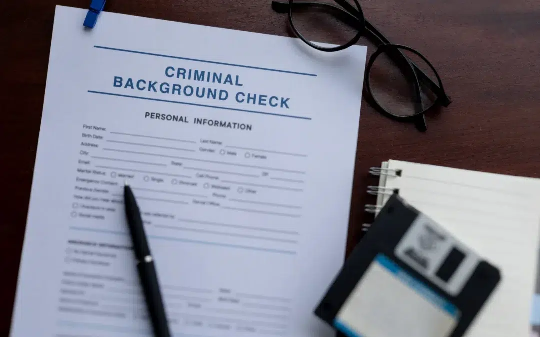 A criminal background check form on a desk, surrounded by a pen, eyeglasses, a notebook, and a floppy disk.