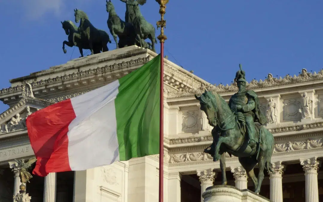 The Italian flag waves in front of the Altare della Patria in Rome, featuring statues of horses and a rider on horseback, with blue sky in the background.