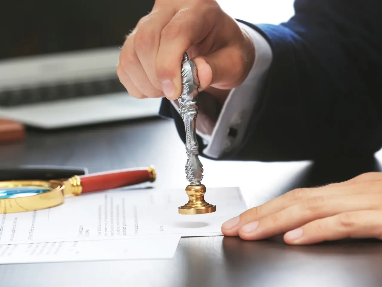 A person in a suit presses a metal stamp onto a document on a desk, with a red pen and magnifying glass nearby.