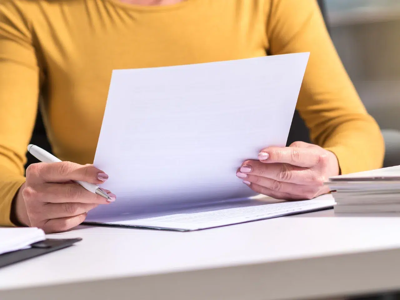 A person wearing a yellow top sits at a desk, holding and reading a sheet of paper with both hands. A pen and a stack of documents are also on the desk.
