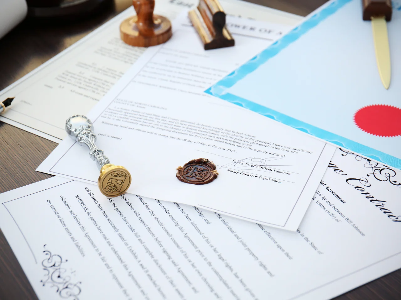 Several official documents and certificates are spread out on a desk, some featuring wax seals and a metallic stamp. A blue-bordered document and a wooden-handled stamp are also visible.