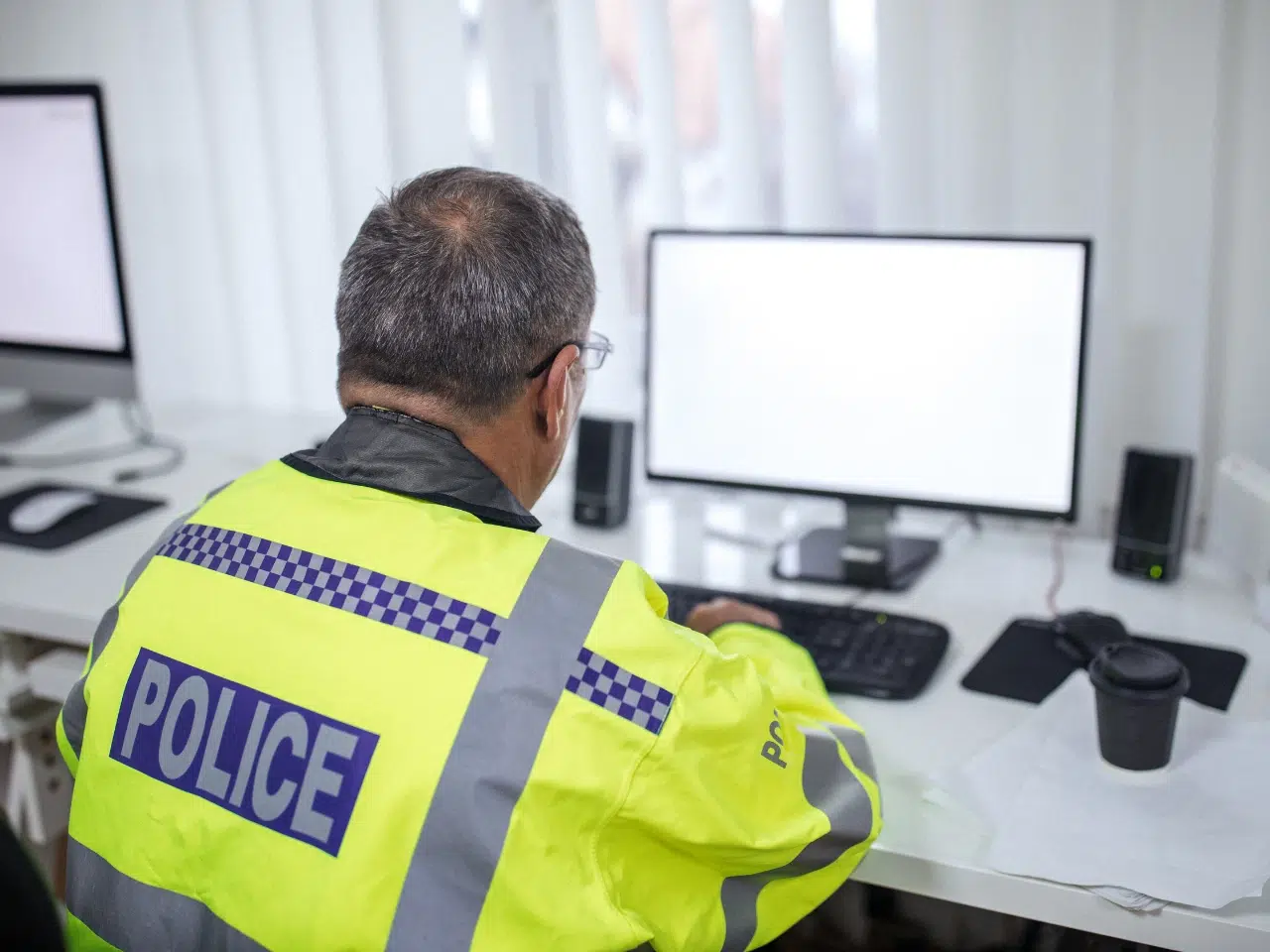 A police officer wearing a high-visibility jacket sits at a desk, working on a computer in an office with white walls and blinds. A coffee cup and papers are on the desk.