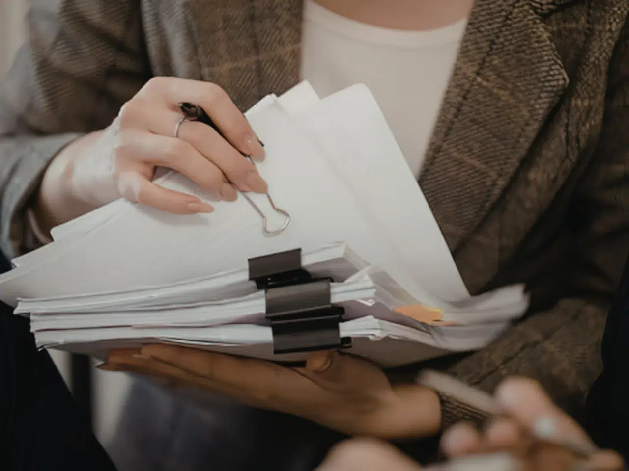 A person wearing a brown plaid blazer holds a large stack of papers clipped together with binder clips, while holding a pen and flipping through the documents.