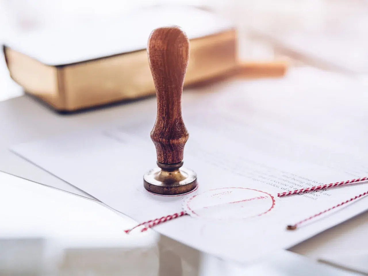 A wooden stamp rests on a stack of official documents with a red wax seal, and a closed book is blurred in the background.