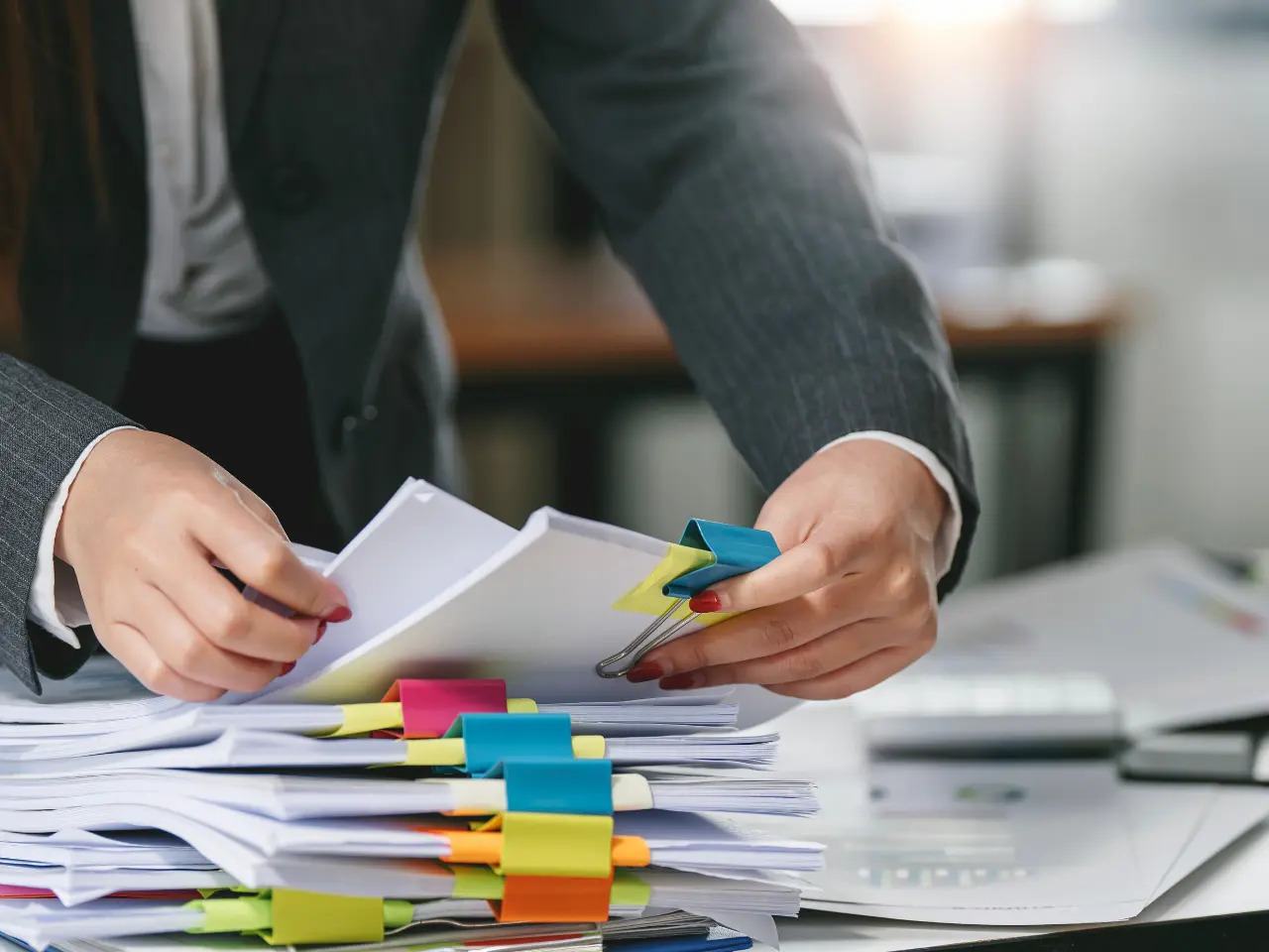 A person in a suit organizes stacks of documents with colorful binder clips and sticky notes on a desk, suggesting office work or paperwork management.