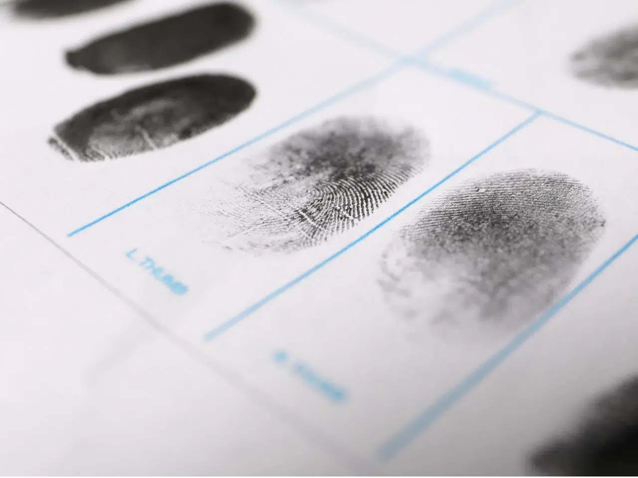 A close-up of black fingerprint impressions on a white paper form, with blue lines separating labeled sections for different fingers. The fingerprint ridges are clearly visible.