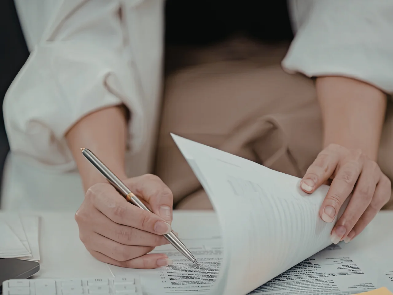 A person holding a pen in one hand and flipping through papers with the other, appearing to review or sign documents at a desk.