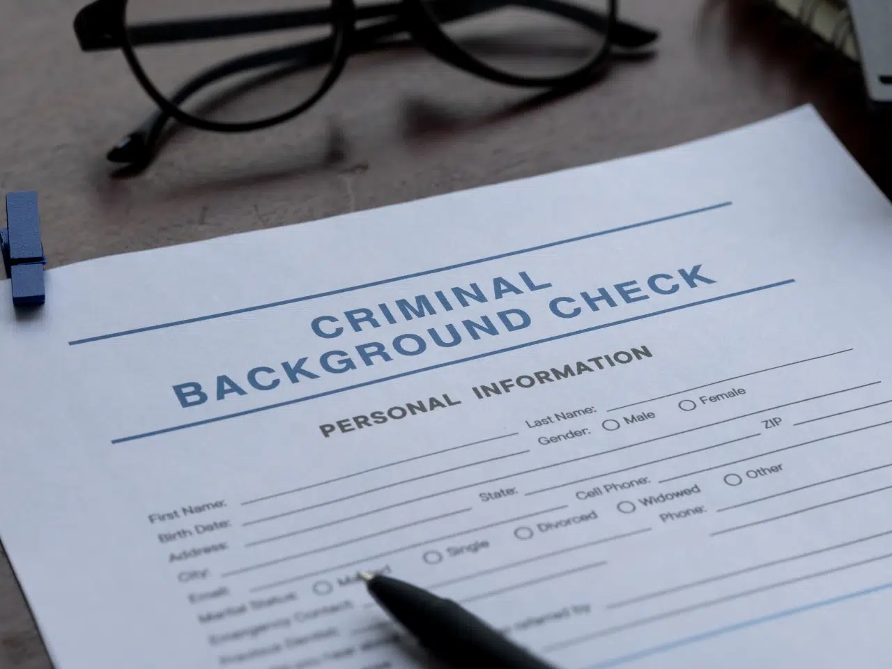 A close-up of a criminal background check form on a desk, with a pen resting on it and a pair of eyeglasses in the background. The form asks for personal information like name, address, and marital status.